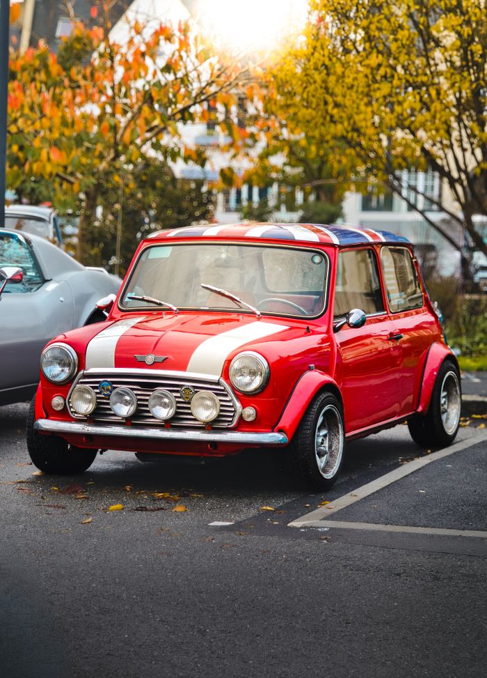 A vintage red Mini Cooper with racing stripes parked outdoors in an autumn setting.